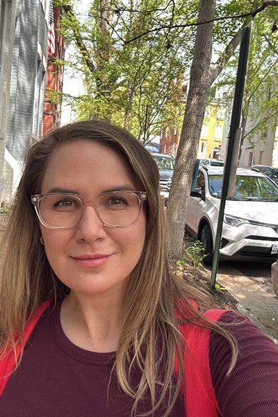 Photo of a woman standing on the sidewalk wearing eyeglasses and a maroon long sleeve shirt.