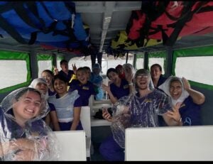 A group of students posing excitedly in a covered boat wearing clear ponchos. 