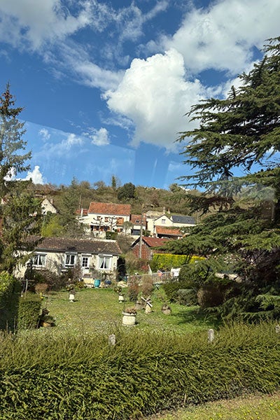 a small French village with cozy houses, red-tiled roofs, and a well-kept garden in the foreground. Tall trees frame the scene, and a brilliant blue sky with large, fluffy clouds stretches overhead.