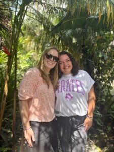 Stephanie and another student standing with trees and greenery in the background. Both are smiling. Stephanie is wearing an ECU t-shirt.