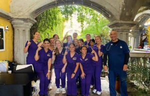 A group of students in purple scrubs standing in front of an archway smiling and holding up a pirate hook finger.