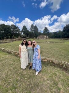 Stephanie and two other students wearing dresses standing in front of a brick ledge on a grassy knoll.