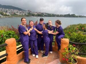 A group of students in purple scrubs standing in front of the river and using their stethoscopes to playfully listen to each other's heartbeat.