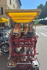 Four women joyfully riding a bicycle together, showcasing teamwork and camaraderie in a sunny outdoor setting. 