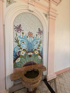 A decorative water fountain fastened on the wall with a floral mural behind it.