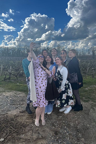 A group of eight smiling women posing for a selfie in a vineyard under a dramatic, cloud-filled sky. They are dressed in stylish, colorful outfits, some with floral patterns, and appear to be enjoying a day out together.
